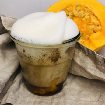 Pumpkin Spiced Latte im Glas. Im Hintergrund liegt ein orangener aufgeschnittener Kürbis. Das Glas steht auf beigen Leinentüchern. Das Getränk ist bräunlich mit weißem Milchschaum. Am Boden setzt sich erkennbar orangenes Kürbispüree ab.