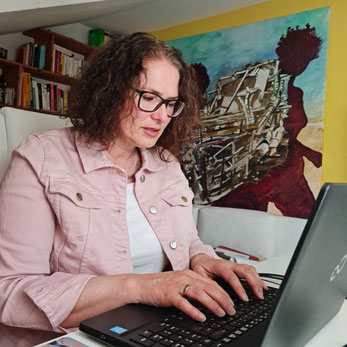 Miriam Pietrangeli sitz am Schreibtisch und schreibt am Laptop. Im Hintergrund sieht man ihr Bild "Sogno"