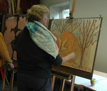 een vrouw tekent met krijt op een groot formaat papier. Op de tekening stat een vrouw op haar knieën met gebogen hoofd, helemaal oranje  en ook de omgeving is warm van kleur.