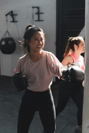 Lächelnde Frau mit Boxhandschuhen im Training am Boxsack, weitere Frau im Hintergrund