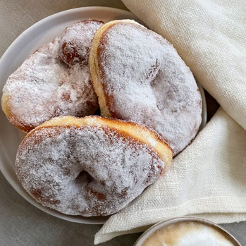 Drei zuckrige Krapfen auf einem weißen Teller, im Anschnitt ein helles Tuch und eine Tasse Cappuccino