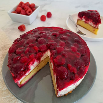 Angeschnittene Himbeertorte mit Cremefüllung und einer Schicht Himbeeren auf einem hellen Biskuitboden, ein Stück auf einem Teller mit Gabel im Hintergrund