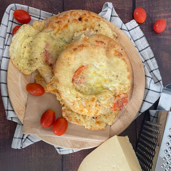 Zwei mit Käse überbackene Tomaten-Käse-Brötchen liegen übereinander auf einem runden Holzbrett, daneben liegen frische Tomate sowie ein Gouda mit einer Käsereibe