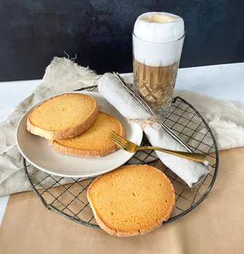 Drei runde Heidesandkekse auf einem weißen Teller und einem Gitter, daneben ein Glas Latte Macchiato mit Milchschaum und eine goldene Gabel auf einer grauen Serviette