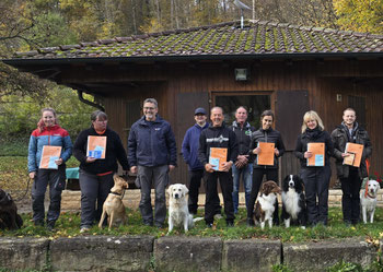 Gruppe von Menschen steht vor einem Holzhaus, jeder hält eine orangefarbene Urkunde, mehrere Hunde sitzen oder stehen vor ihnen auf einer Wiese mit herbstlichen Bäumen im Hintergrund.