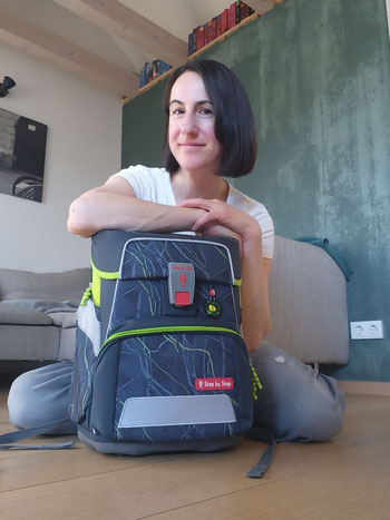 Michaela Dauscher sitzt auf dem Boden, vor ihr eine Schultasche, sie stützt die Arme darauf ab und lächelt. Im Hintergrund eine grüne Wand und ein Sofa.
