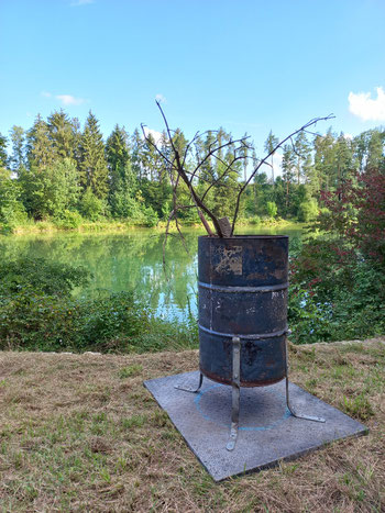 Eine große Feuertonne steht vor einem Fischweiher, Äste ragen heraus