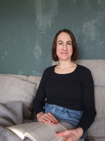Michaela Dauscher sitzt auf einem Sofa, vor einer grünen Wand. Lächelt, ist zugedeckt und hat ein Buch auf ihrem Schoß. Sie trägt ein schwarzes Shirt, ihre Haare sind braun und schulterlang.