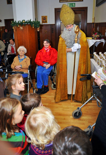 Der Nikolaus mit seiner goldenen Mitra und seinem goldenen Bischofsstab ist umgeben von älteren Menschen im Rollstuhl und von jungen Kindern, die erwartungsvoll auf den Schokonikolaus schauen.