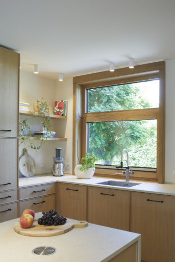Vertikales Schiebefenster aus Holz in moderner Küche mit Blick ins Grüne – geöffneter Fensterflügel über Arbeitsplatte mit Obst und Saftglas.