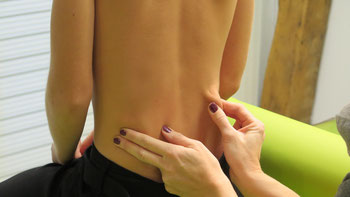 Frau Tollas massiert den Rücken eines Patienten, welcher aufrecht auf einer Massageliege sitzt. Hierzu wendet Sie die Behandlung der Bindegewebsmassage an.