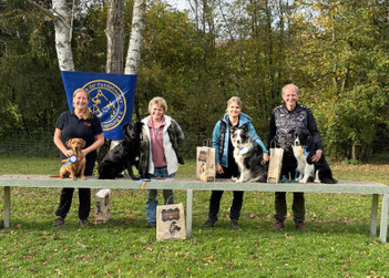 Vier Personen mit jeweils einem Hund unterschiedlicher Rassen stehen hinter einer Bank im Freien, auf der mehrere Papiertüten mit Hundemotiven stehen.