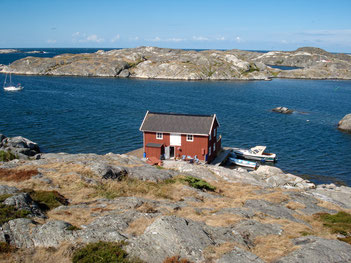 Ein rotes Holzhaus mit weißen Fenstern steht auf einer Schäreninsel, davor liegen zwei Boote 