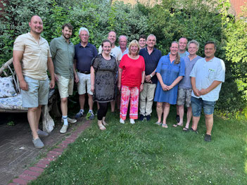 Insgesamt 13 Männer und Frauen stehen im Freien mit sommerlichen Klamotten und posieren für ein Gruppenfoto.
