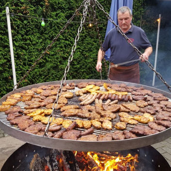 Großer runder Grill mit zahlreichen Steaks, Würstchen und Hähnchenstücken, darüber hängt eine Kette, im Hintergrund steht eine Person mit dunkelblauem Hemd und brauner Schürze