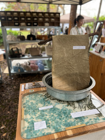 山口アーツ&クラフツ２０２６にてD -29ブースにて出店する、帆布バッグのお店Cargo Ship。小豆島のオリーブの葉と讃岐山脈の湧き水を掛け合わせて作るオリーブ帆布がディスプレイされていて、ガラステーブルの下には、シーグラスとオリーブの葉が敷き詰められています。ブースの奥には横長ボストンバッグducksやお仕事トートバッグなどが並んでいます。