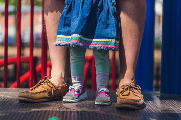 Füße eines Mannes in Lederschuhen, dazwischen Füße eines Kindes in Sandalen. ZUsammen stehen Vater und Tochter auf einem Spielplatz.