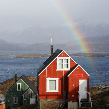 The picture shows a small red building of the hotel "The Red House". A rainbow, the water surface of a fjord and mountains are recognisable in the background.