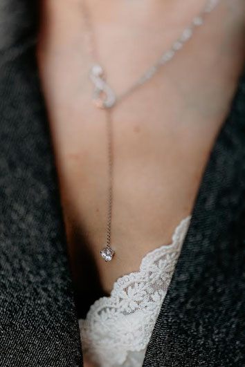 Close-up of a silver pendant with a clear stone on a delicate chain, worn over soft lace and grey fabric. Elegant and feminine detail shot.
