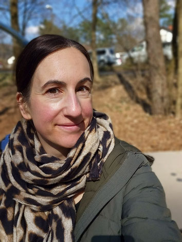 Selfie von Michaela Dauscher, trägt die Haare nach hinten gebunden, einen Schal mit Leo Muster und eine grüne Jacke. Hinter ihr sind Bäume zu erkennen.