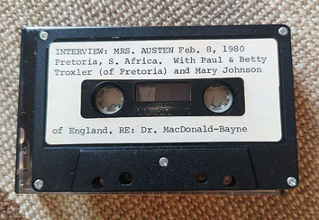 Es ist eine schwarze Musik-Cassette mit einem weißen Aufkleber zu sehen, auf dem geschrieben steht: INTERVIEW: MRS. Austen Feb. 8, 1980 Pretoria, S. Africa. With Paul & Betty Troxler (of Pretoria) and Mary Johnson of England. RE: Dr. MacDonald-Bayne