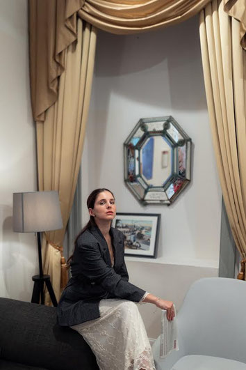 Woman in a grey blazer and white lace dress sitting in an elegantly decorated room. Heavy curtains and a decorative mirror behind her create a calm and stylish atmosphere.