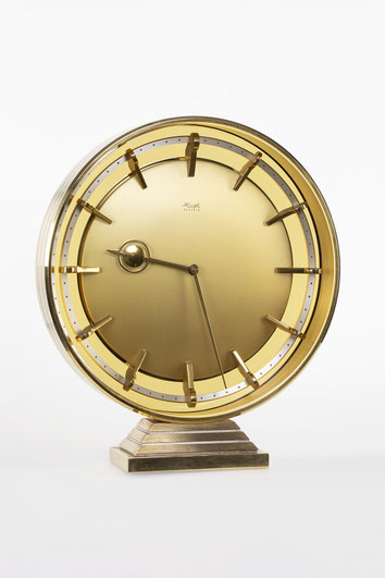 goldene, kreisrundes Tischuhr auf einem kleinen, mehrstufigen Sockel. Das Zifferblatt ist Vorder- und Rückseitig verglast. Das hintere Glas ist gelblich eingefärbt.