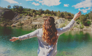 Woman standing with raised arms by lake to be happier