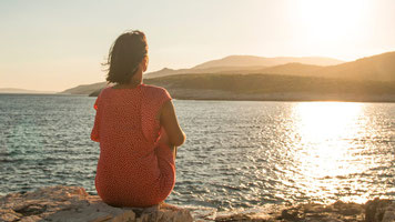 Eine Frau sitzt am Ufer und schaut auf die Berge und das Meer