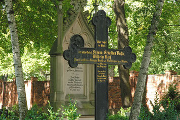 Passend zur Bach-Genealogie sieht man ein Kreuz verziert in schwarz mit einem Grabstein im Hintergrund. Es steht auf einem Friedhof unter Bäumen. Hinten sieht man die Sonne auf einigen Gräbern. Das Kreuz trägt eine Inschrift zur Bach-Genealogie.