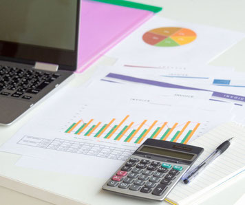 Plan de travail de bureau, avec un ordinateur portable ouvert sur la gauche, et des feuilles avec des graphiques sur la droite, sur lesquelles sont posés une calculatrice et un stylo.
