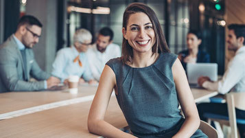 Ein professionelles, modernes Büro-Setting mit einer selbstbewussten, lächelnden Frau im Vordergrund, die Authentizität und Führungsstärke ausstrahlt. Im Hintergrund sind mehrere Teammitglieder in einer Besprechung, was ein dynamisches und kollaboratives 
