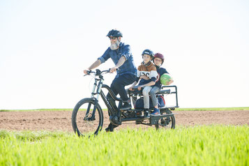 Mann mit Bart fährt mit dem XCYC Pickup Life und seinen zwei Kindern über ein Feld