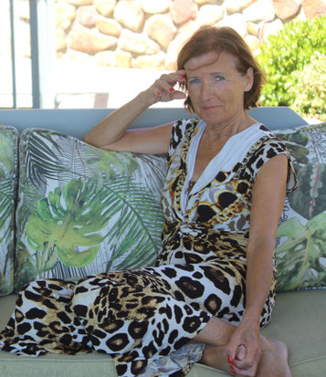 Margit Königsecker, Coach & Trainerin - sitzt sommerlich gekleidet auf einem gemusterten Sofa, rechte Hand seitlich an der Stirn - sieht in die Kamera