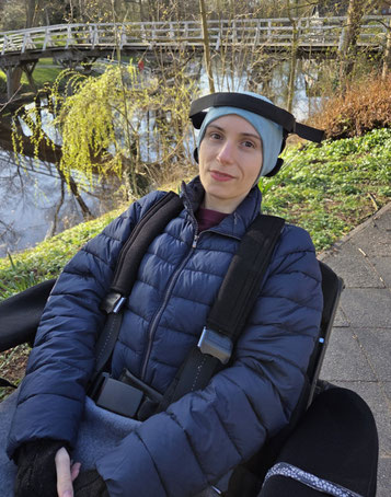 Johanna im E-Rollstuhl, sitze seitlich zur Kamera und lächel. (2026)