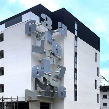 Public wall sculpture by Paolo Grassi in Massagno, Switzerland, corten numerals integrated into residential facade.