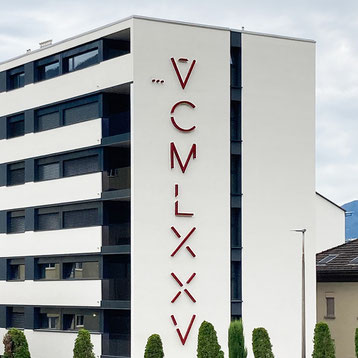 Vertical wall sculpture by Paolo Grassi titled VCMLXXV, red numeric installation on modern white facade in Locarno.