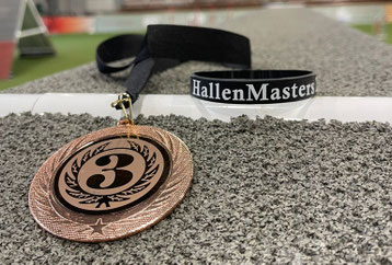 Bronzene Medaille mit der Zahl 3 und einem schwarzen Band mit dem Schriftzug 'HallenMasters' liegt auf grauem Teppichboden