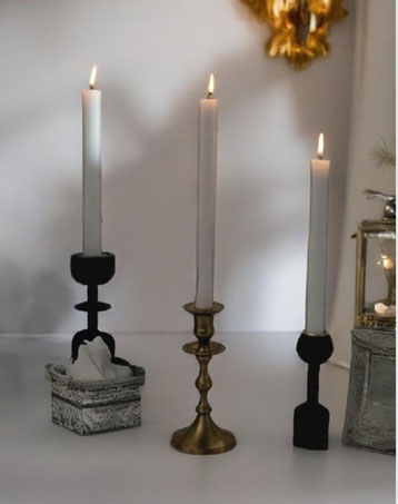 Drei brennende Stabkerzen in unterschiedlich geformten Kerzenhaltern aus Schwarz und Gold auf einer hellen Oberfläche. Die Szene wirkt warm, minimalistisch und stilvoll.
