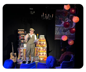 Live-Foto von Thommi Baake auf der Bühne in Hannover: lesend vor stimmungsvollem Bühnenbild, Gitarre im Hintergrund