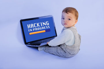 Infant boy sitting with laptop computer resting on his legs and looking over shoulder at the camera. The screen has a yellow progress bar and the words "Hacking in Progress". 