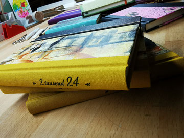 Zu sehen ist ein Buch (Kalender für 2024). Der Rücken trägt ein gelbes Gewebe mit einer Jahreszahl-Prägung in schwarz. Der Vorderdeckel ist mit einem selbstgestaltetem Unikatpapier überzogen. Im Hintergrund liegt eine Schere und Falzbein sowie eine Binde.