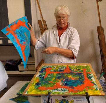 Een vrouw met wit haar en een witte bloes aan, heeft net een afdruk gemaakt met verf op een vel op een schilderij in de vorm van een oranje kader. Ze kijkt verrast naar het resultaat.