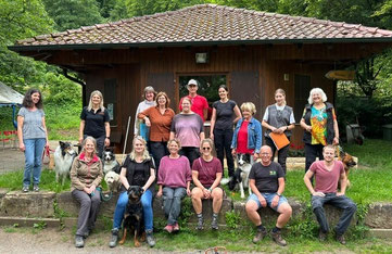 Gruppe von Menschen vor einem kleinen Holzhaus mit Ziegeldach, einige sitzen auf einer niedrigen Mauer, andere stehen, mehrere Hunde sind dabei.