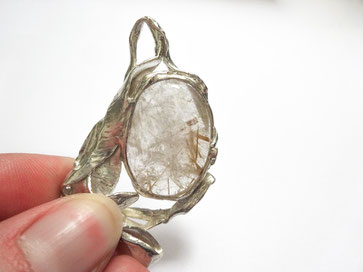 Un gros pendentif en argent avec une belle pierre transparente blanche aux inclusions dorées est exposée sur fond blanc.