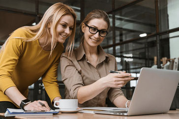 Zwei Frauen tauschen sich im Büro über Ihre Ausarbeitungen aus. Eine Frau mit Brille und braunen Haaren sitzt vor dem Laptop. Eine Frau mit langen blonden Haaren steht und beugt sich zu ihr herunter. Beide lächeln in in den Laptop. 