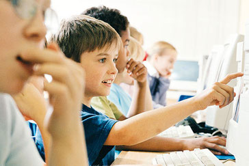 Kinder sitzen nebeneinander an Computern, ein Junge zeigt auf den Bildschirm eines PCs