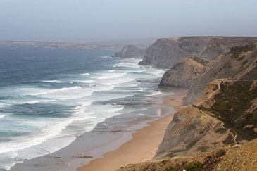 Blick von oben auf die Costa Vicentina mit hohen Felsklippen und dunkelgelbem Strand, das Wasser ist weiß von heranrollenden Wellen