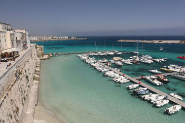 Hafen von Otranto mit türkisfarbenem Wasser und mit einer Reihe kleiner Boote. Auf der linken Seite die Stadtmauer