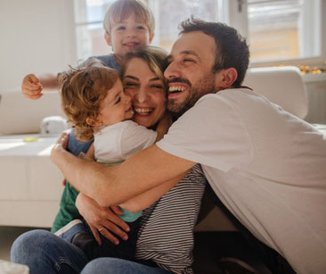 Ein Mann und eine Frau mit 2 kleinen Kindern, die sich alle umarmen und lachen - symbolisch für tiefes Vertrauen, sichere Bindung und emotionale Freiheit - mein schützling